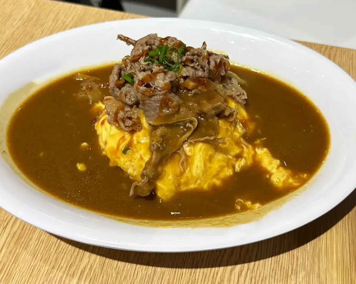 A white plate with Japanese curry rice topped with a fluffy omelette, thinly sliced beef, and garnished with green onions, served on a wooden table.