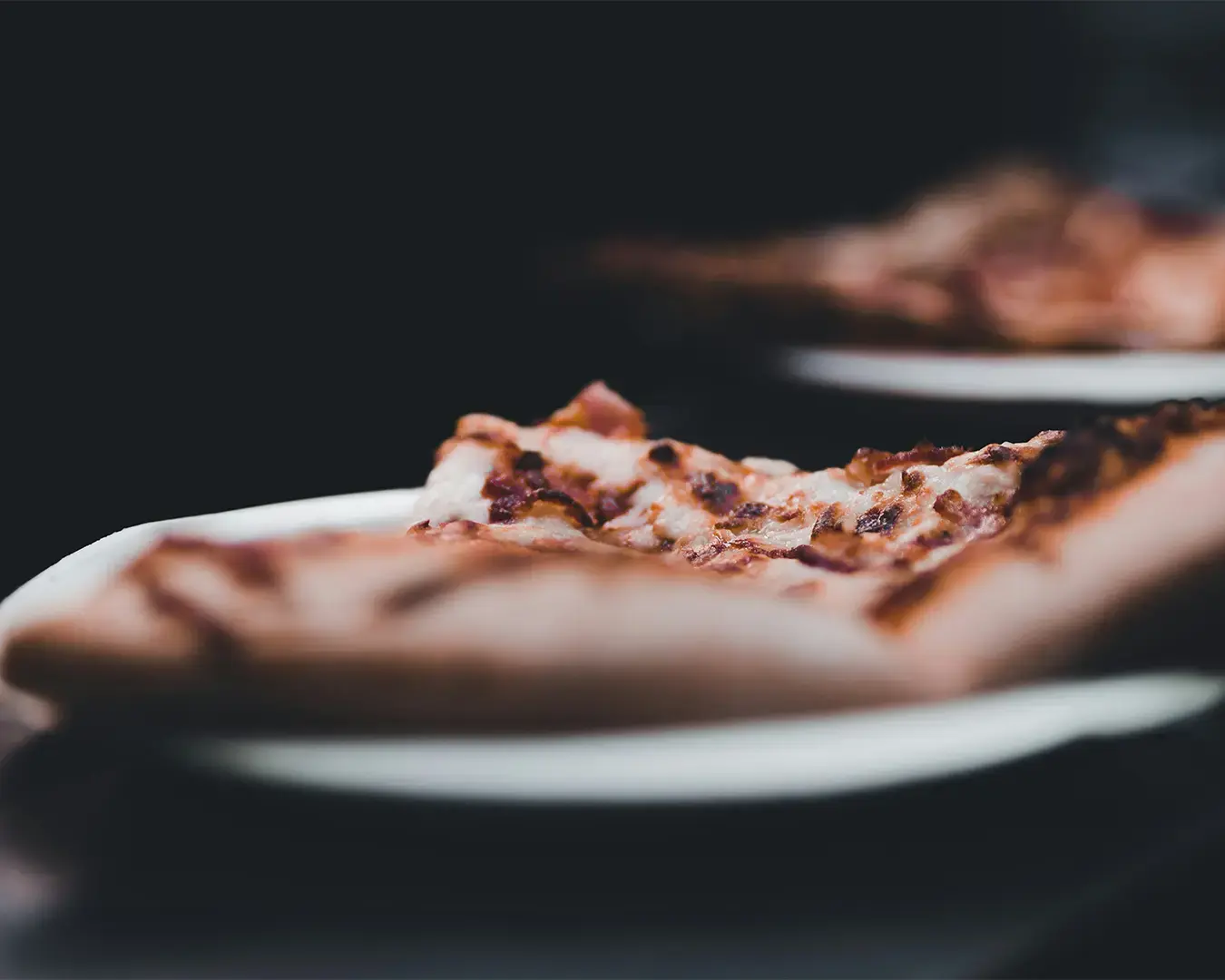 Close-up of a slice of pizza on a white plate against a dark background. The pizza has a crispy crust and visible toppings like bacon.
