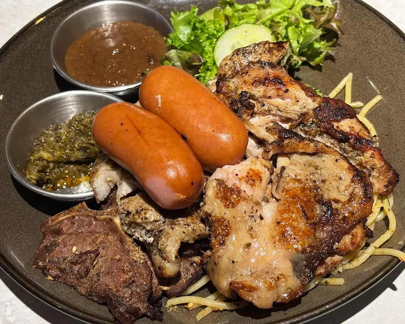 A plate with grilled chicken, sausages, and steak, accompanied by pesto and brown sauce. Garnished with lettuce, cucumber, and spaghetti strands.