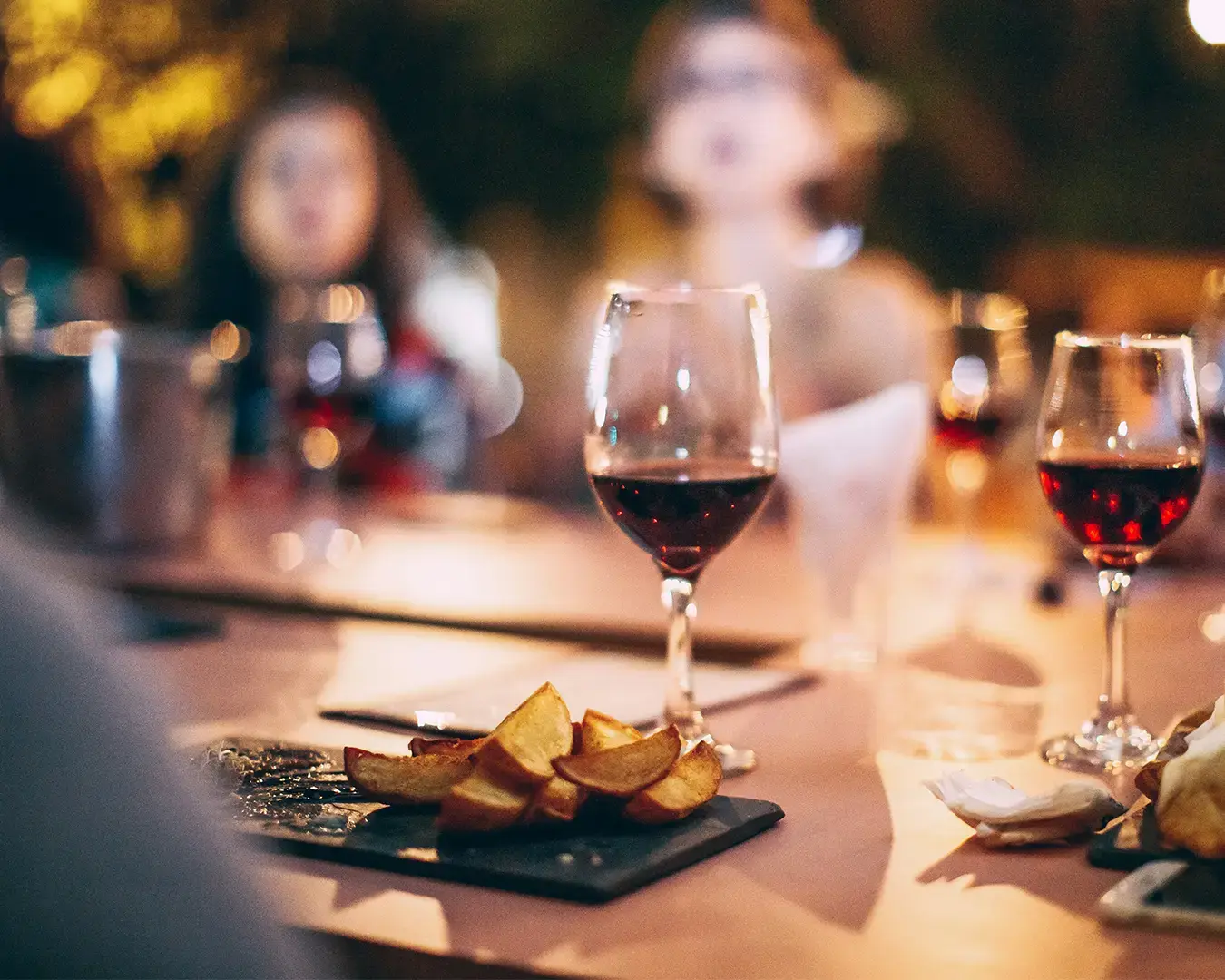 A cozy dinner setting with a focus on red wine glasses and a slate plate of potato wedges. Blurred background shows people enjoying the warm atmosphere.