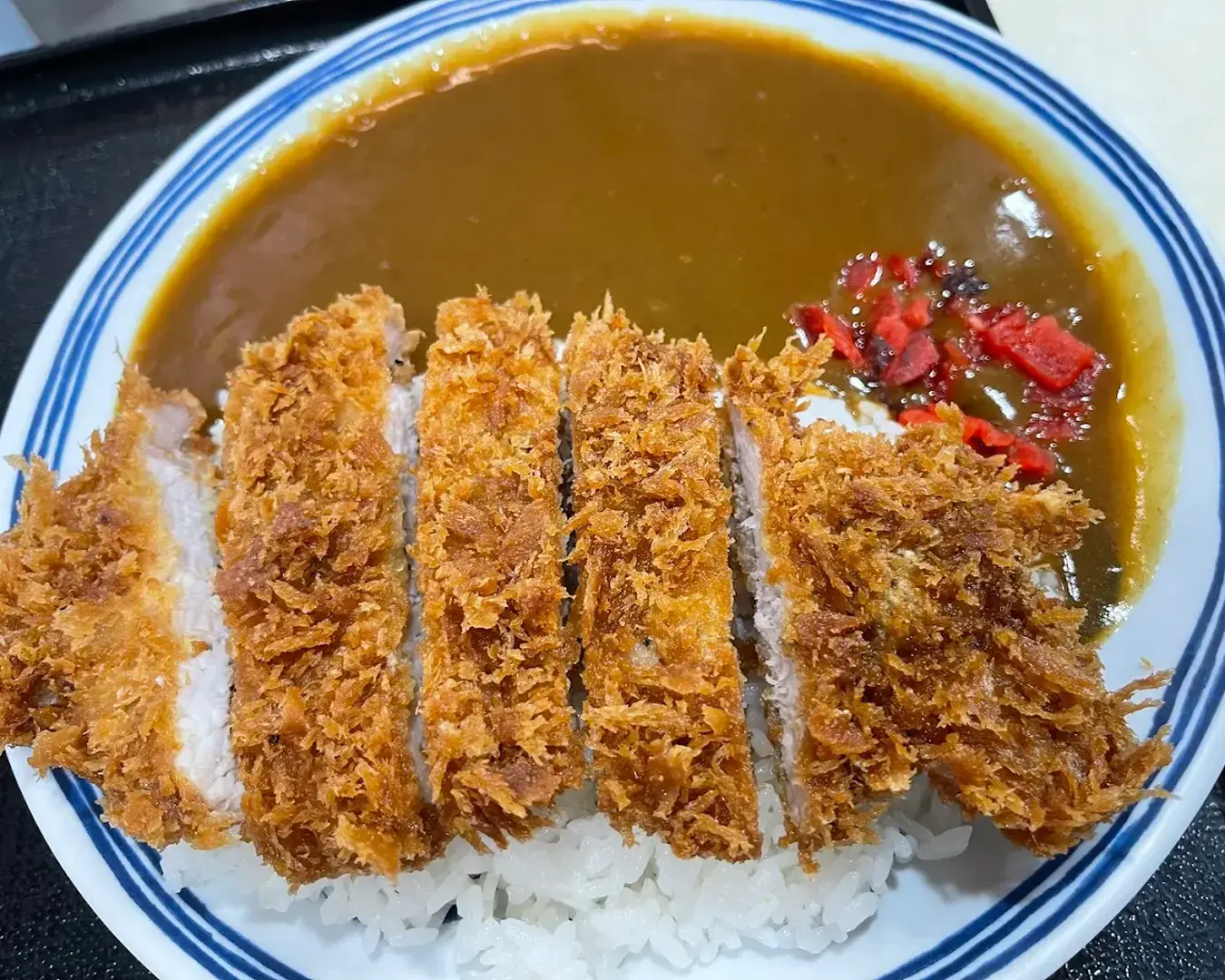 A plate of Japanese curry rice topped with crispy, golden-brown pork cutlets. Rich brown curry sauce and red pickled ginger add vibrant contrast.