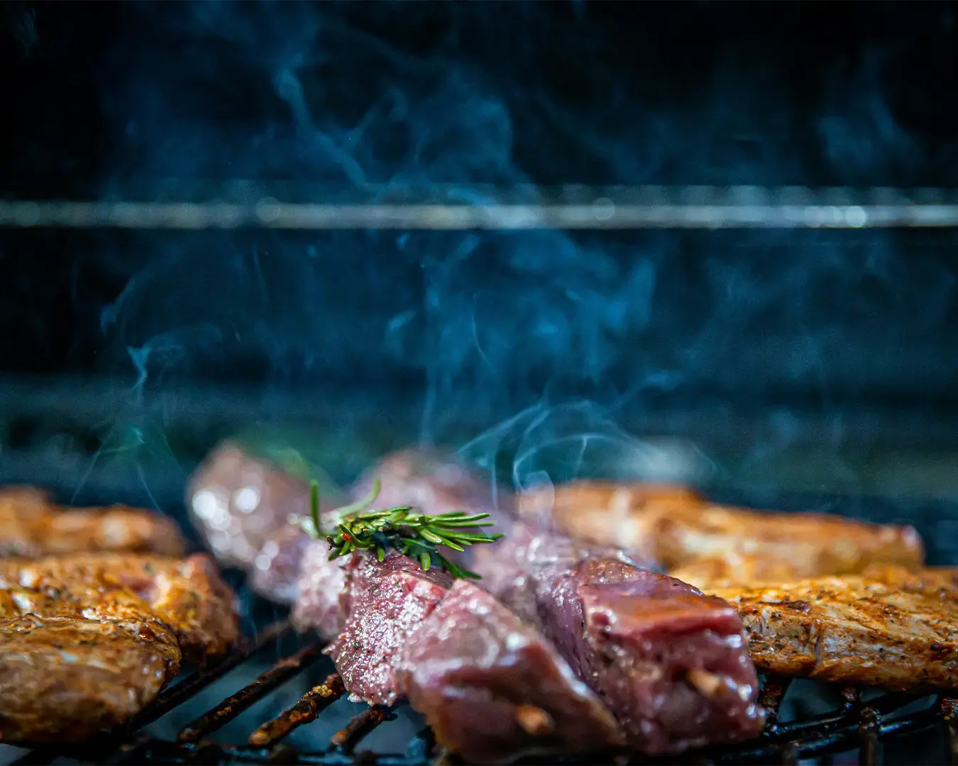 Steaks sizzling on a grill with smoke swirling above. Fresh rosemary adorns the meat, enhancing the savory and aromatic atmosphere of a barbecue.