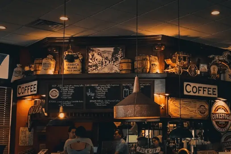 Warmly lit cafe ordering counter decorated with rustic Western elements, including burlap coffee bean sacks, small wooden barrels, and a miniature stagecoach sitting above handwritten chalkboard menus.