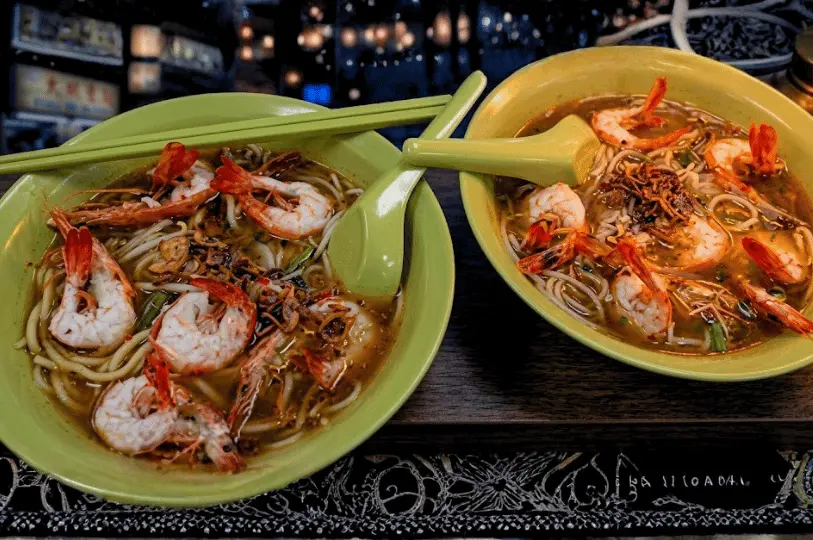 Close-up food photography of two green bowls of traditional Singaporean prawn noodles, showcasing vibrant red prawns and rich broth set against a dark, moody midnight ambiance.