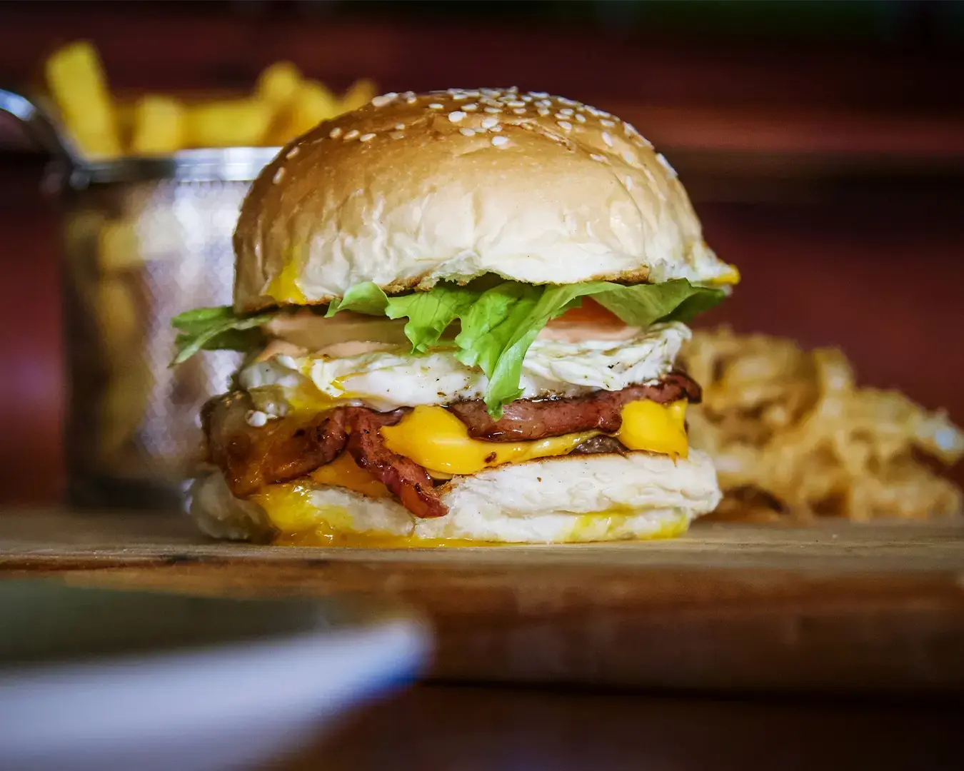 Close-up of a juicy burger with sesame seed bun, crispy bacon, lettuce, melted cheese, and fried egg on a wooden board; fries in the background.