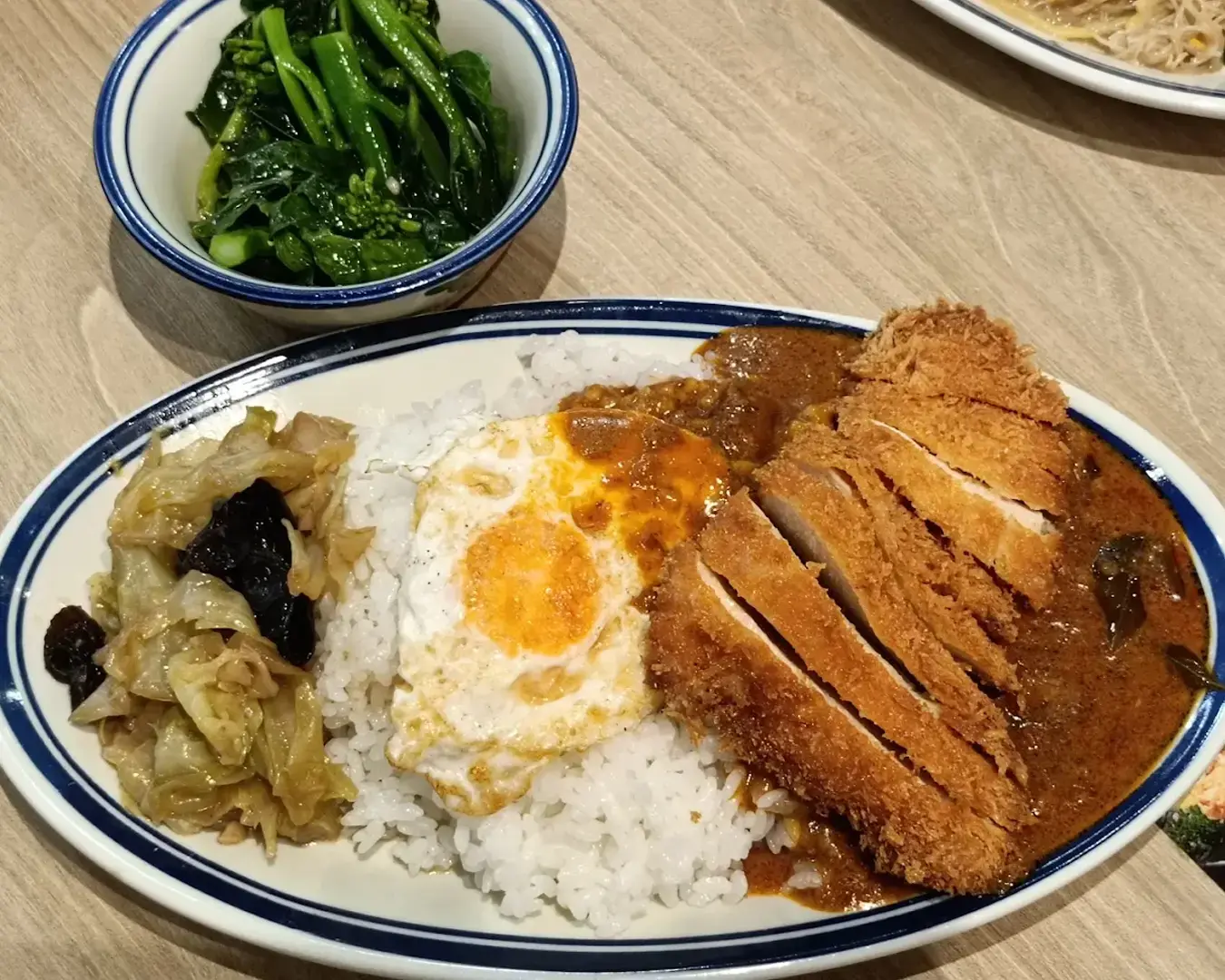 A plate of rice topped with fried egg and curry sauce, accompanied by sliced breaded meat and cabbage. A side of leafy greens in a small bowl.