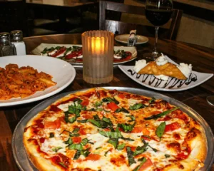 A cozy dinner setting with a pizza topped with fresh basil, pasta, caprese salad, and a dessert plate featuring a cannoli. A candle glows warmly.