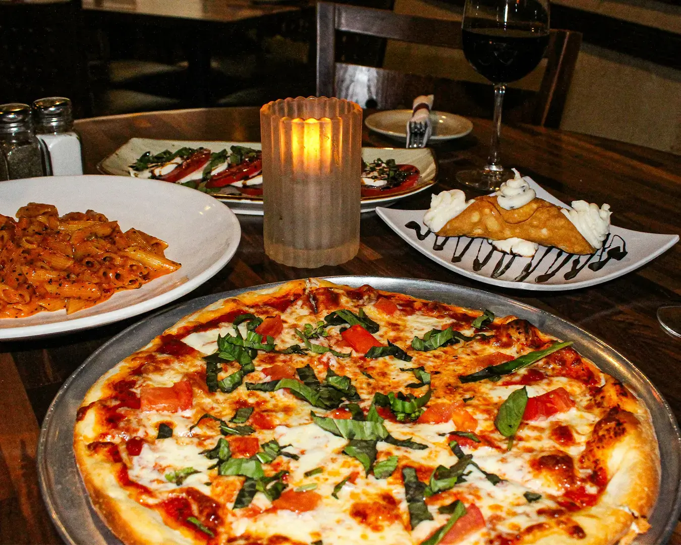 A cozy dinner setting with a pizza topped with fresh basil, pasta, caprese salad, and a dessert plate featuring a cannoli. A candle glows warmly.