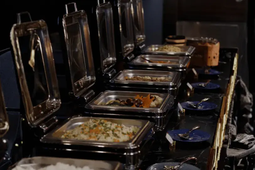 A dimly lit buffet station featuring a row of open stainless steel chafing dishes filled with hot foods like rice and mixed vegetables, lined up on a black marble counter.