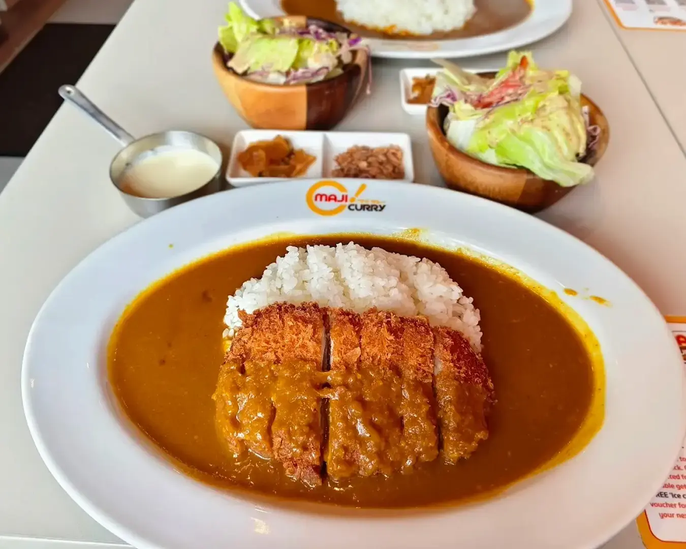 A plate of katsu curry features breaded cutlet and white rice smothered in golden curry sauce. Sides include salad, pickles, and a creamy sauce.