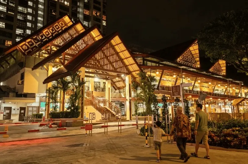Architectural night photography of Geylang Serai Market, highlighting its distinctive traditional peaked roofs glowing with warm golden lights against the dark sky as passersby approach the entrance.