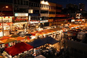 An elevated view of a vibrant night market street bustling with crowds, lined with rows of colorful illuminated tents and glowing storefront signs under the warm night sky.