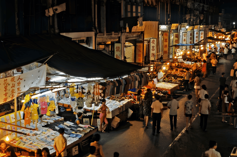 A realistic, atmospheric street scene of a traditional night market where pedestrians stroll past clothing stalls under canvas awnings and buildings displaying vintage Chinese signage, all illuminated by warm, glowing lights.