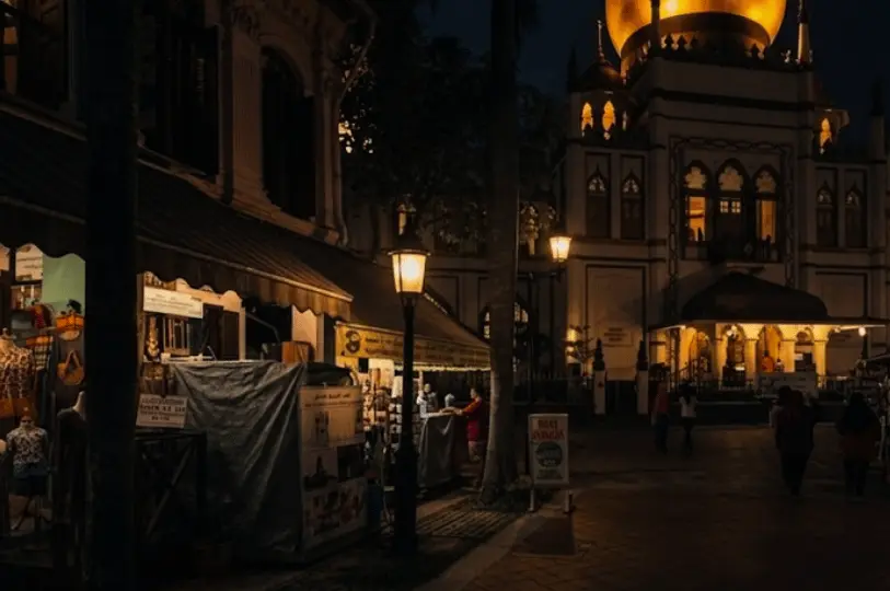 A night view of the Sultan Mosque in Kampong Glam, Singapore, with its golden dome glowing against the dark sky, framed by street lamps and traditional shophouses.