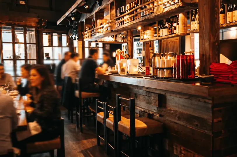 Warm, dimly lit interior of a Japanese izakaya showing a rustic wooden bar counter lined with seating, overhead shelves stocked with liquor bottles, and blurred diners in the background.