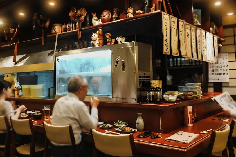 Diners seated at a red bar counter inside a cozy Japanese izakaya, featuring a glass seafood display case and overhead shelves decorated with traditional figurines like Daruma dolls.