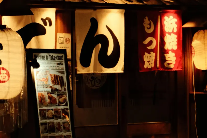 Close-up of an izakaya storefront at night, featuring traditional fabric noren curtains with large Japanese characters, glowing paper lanterns, and an illuminated menu board showing grilled skewers.