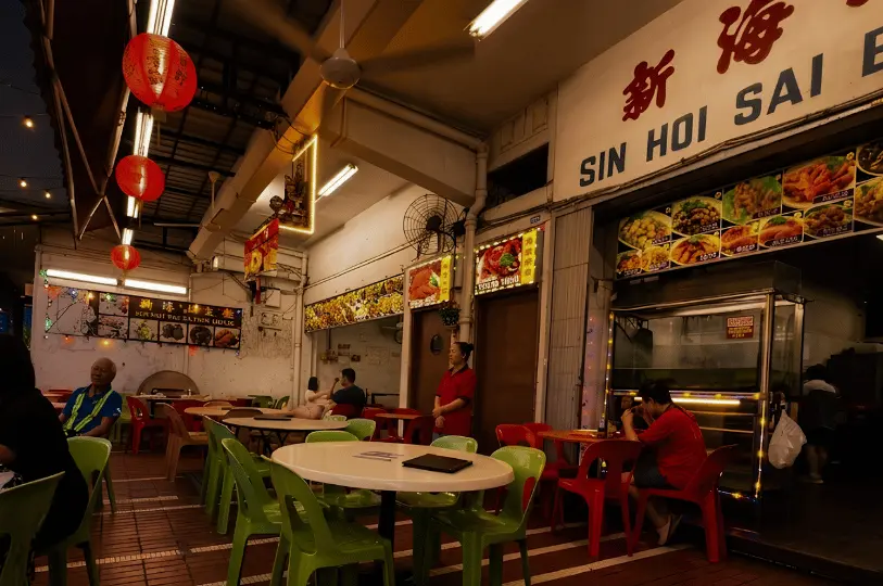Diners seated at round tables with red and green plastic chairs under the bright lights and hanging red lanterns of the open-air Sin Hoi Sai Eating House.
