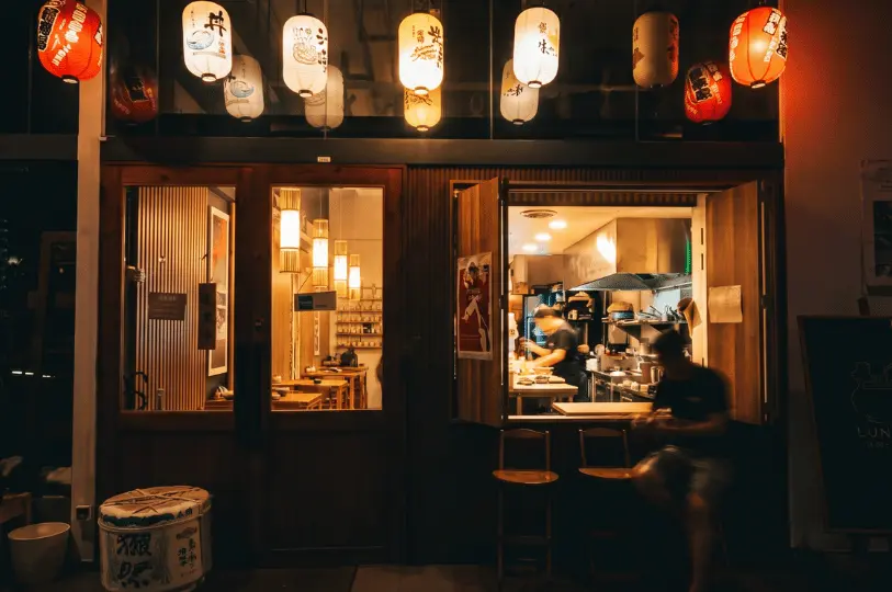 The exterior view of a warmly lit izakaya at night, showing a chef cooking inside through an open window, with glowing paper lanterns hanging above the entrance.