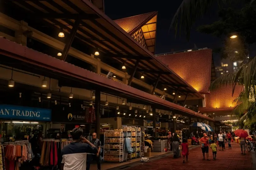Pedestrians browsing shops and strolling along a brick-paved walkway under the distinctive, warmly lit traditional Malay architecture of the Geylang Serai market area at night.