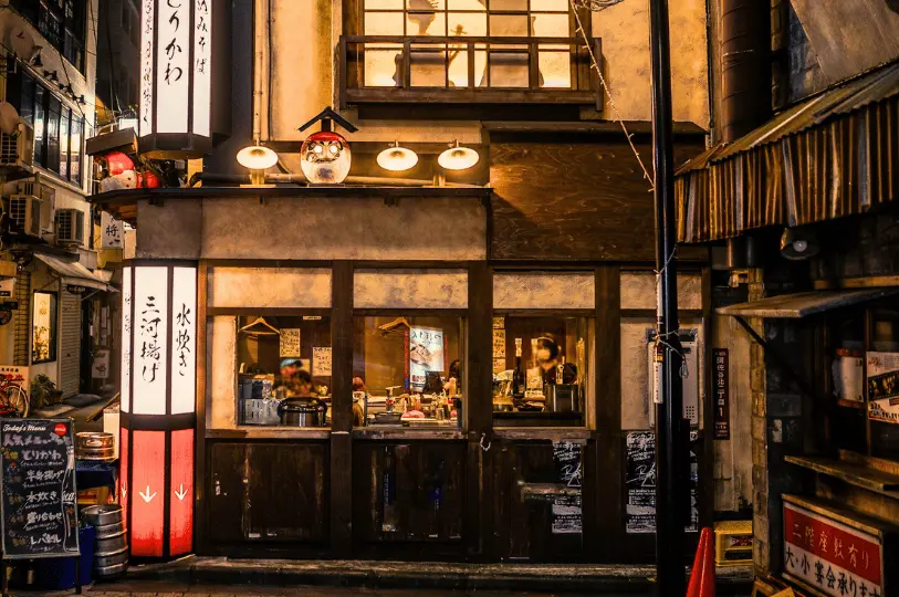 Exterior night view of a traditional-style Japanese izakaya illuminated by glowing lanterns and vertical signs, with patrons visible dining inside the cozy ground-floor windows.