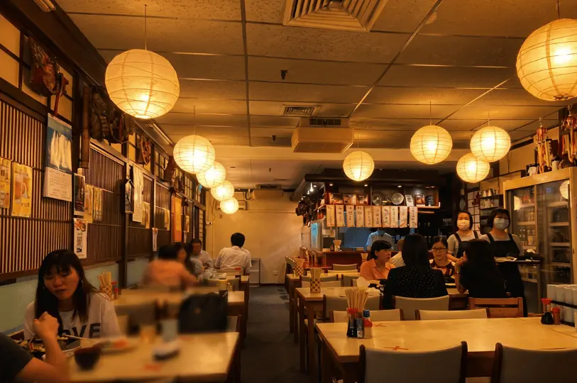 The cozy interior of a bustling Singapore izakaya illuminated entirely by warm, low-hanging paper lanterns above diners at wooden tables.