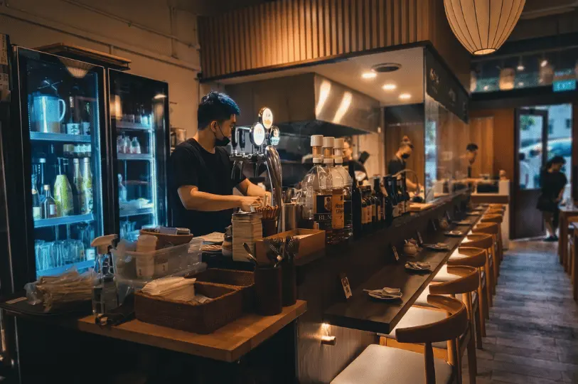 A bartender wearing a face mask prepares drinks behind a wooden counter at a Singapore izakaya, with glowing blue drink refrigerators and neatly arranged seating visible.