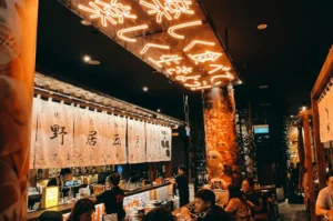 Lively interior of a late-night izakaya featuring bright orange neon Japanese characters on the ceiling, traditional white noren curtains over the bar, and patrons enjoying drinks.