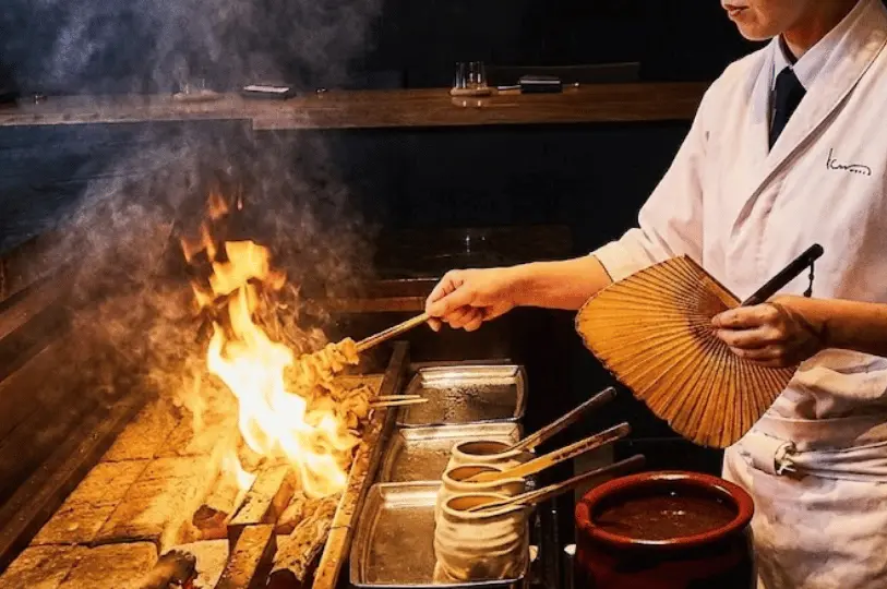 A dramatic low-light action shot of an izakaya chef using a traditional fan to control bright, leaping flames over a charcoal yakitori grill.