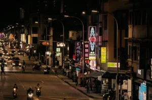 A bustling nighttime street scene in Geylang, Singapore, illuminated by overhead streetlights and bright neon restaurant signs with Chinese characters, as cars and scooters navigate the road.