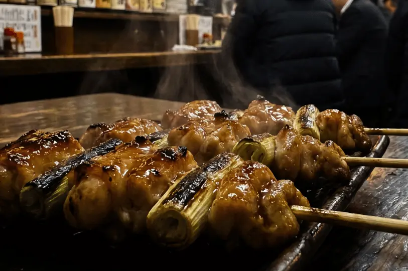 A close-up food photography shot of steaming, glossy yakitori chicken skewers resting on a dark surface under warm, directional izakaya lighting.