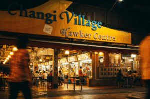 A vibrant nighttime view of the Changi Village Hawker Centre. The large yellow entrance sign is brightly illuminated above numerous glowing food stalls. Patrons are seated at outdoor tables enjoying late-night meals, while blurred pedestrians walk past in the foreground, capturing the lively midnight atmosphere.