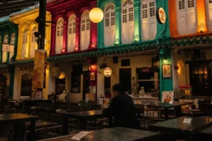 An outdoor night scene of colorful heritage shophouses in Singapore housing izakayas, illuminated by warm yellow street lamps and glowing shop signs.