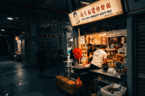 A brightly illuminated hawker stall named "Nan Ling Hokkien Kien Mee" stands out against the dark, quiet surroundings of a dimly lit food center. Inside the glowing stall, a solitary worker in a white t-shirt is busy preparing food among stacked bowls and equipment, capturing the peaceful, anticipatory atmosphere of a hawker center before the bustling crowds arrive.