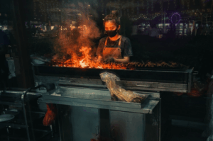 A street food vendor wearing a black face mask and apron focuses intently on grilling a long row of skewers over a blazing charcoal grill at night. Bright orange flames and thick smoke rise from the grill, illuminating the vendor against the dark, moody background of the night market.