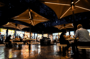 A nighttime scene at a bustling outdoor food centre under a starry sky. Diners sit at circular tables beneath large, modern umbrella canopies lined with warm, glowing lights. The ambient light reflects off the textured stone pavement, while brightly lit food stalls in the background display glowing signs for "LAKSA" and "SATAY", creating a moody, illuminated atmosphere.