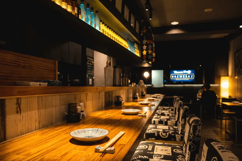 A cozy, warmly lit izakaya interior showing a long wooden dining counter meticulously set with ceramic plates and chopsticks, with shelves of colorful bottles overhead and patrons dining quietly in the background.