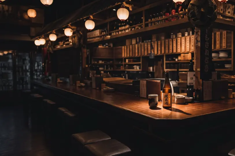 The atmospheric, dimly lit interior of an izakaya, focusing on a long, polished wooden bar counter where a solitary bottle of sake and a cup sit under a warm spotlight, with shelves of bottles in the background.
