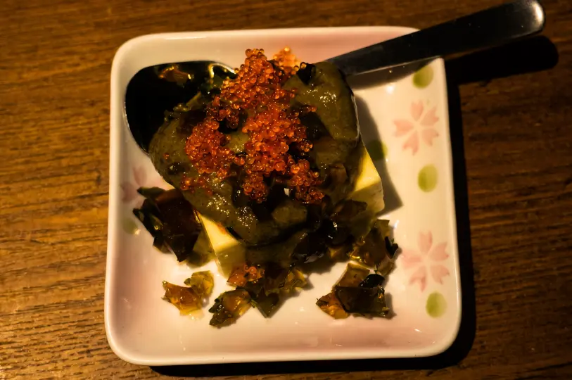 A square ceramic plate with cherry blossom motifs holding a delicate appetizer of tofu topped with a dark, savory paste, shimmering jelly bits, and a mound of vibrant orange fish roe.