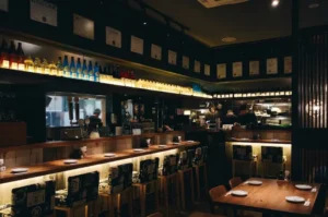 A warmly lit interior of a Japanese izakaya featuring a wooden bar counter, cushioned stools, shelves lined with sake bottles, and chefs preparing food in the open kitchen.