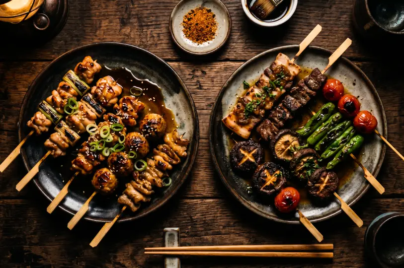 Overhead view of a rustic wooden table featuring two ceramic plates loaded with freshly grilled yakitori skewers, including glazed chicken, shiitake mushrooms, blistered shishito peppers, and charred cherry tomatoes.