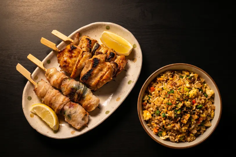 A white plate with four freshly grilled Japanese meat skewers and a lemon wedge, placed next to a bowl of golden-brown, savory fried rice.