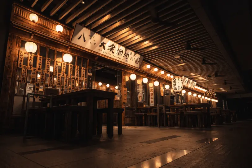 The warm, inviting exterior of a late-night izakaya, featuring a rustic wooden facade covered in Japanese menus and illuminated by rows of paper lanterns above empty wooden outdoor tables.