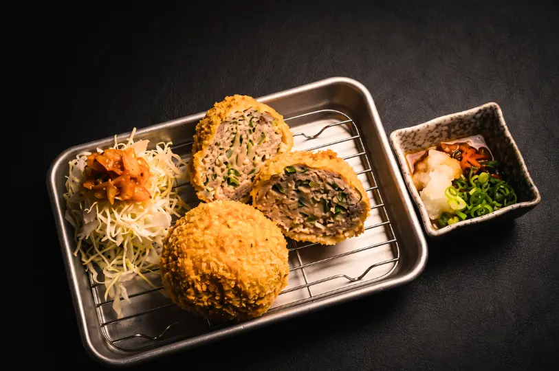 Crispy, golden-brown menchi katsu (fried minced meat patties) served on a metal wire rack with shredded cabbage and dipping sauce; one patty is cut open to reveal a juicy, vegetable-studded meat filling.