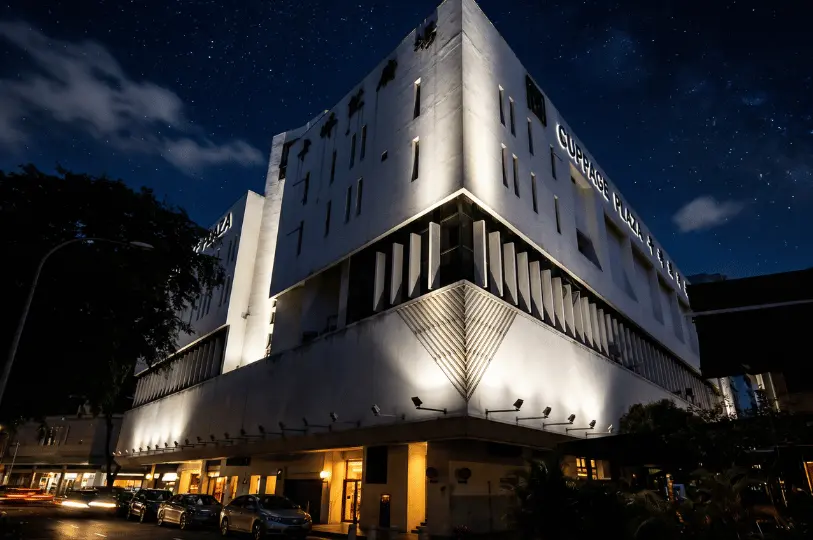 Low-angle exterior architectural shot of the stark white Cuppage Plaza building at night, illuminated by dramatic uplighting that creates deep shadows along its facade against a starry sky.