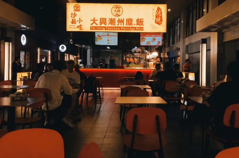 The bright, glowing storefront sign of Tai Heng Teochew Porridge Rice with several patrons seated at tables with orange chairs eating late-night meals.