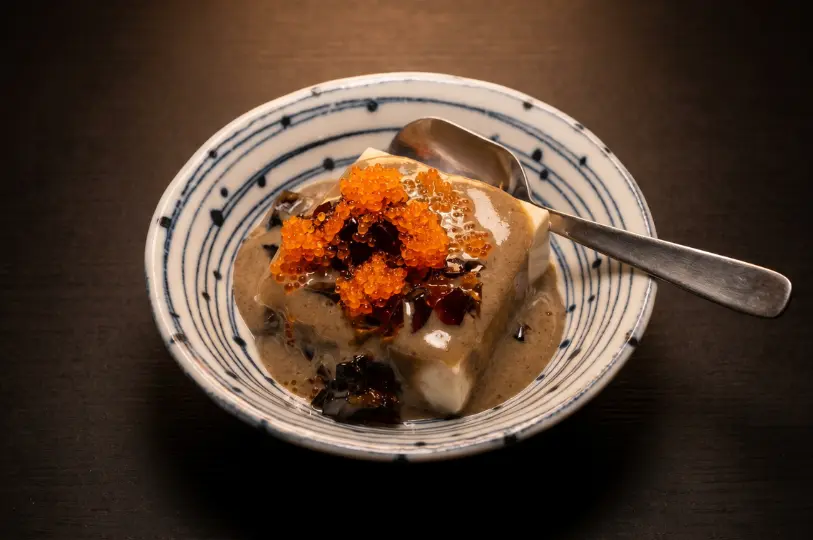 A close-up of a Japanese appetizer in a round patterned bowl, consisting of a block of tofu covered in a rich, dark grey sauce and topped with bright orange fish roe and jelly pieces.