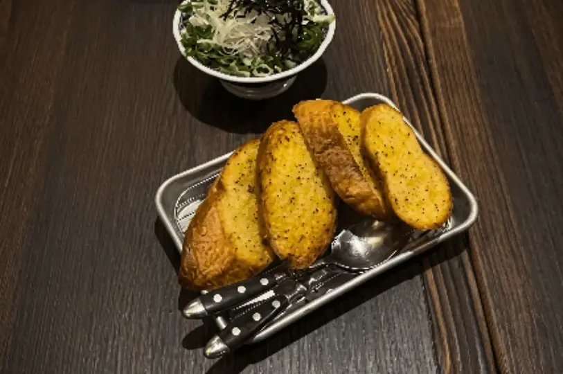 A top-down view of a dark wooden table featuring a small metal tray holding four thick slices of toasted, seasoned bread resting on top of black-handled utensils. Above the tray is a small bowl filled with a fresh garnish of shredded cabbage, green onions, and strips of nori seaweed.