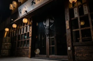 Exterior of an authentic-looking izakaya at night, featuring a dark wooden facade, warm glowing paper lanterns, sliding doors, and stacked sake barrels near the entrance.