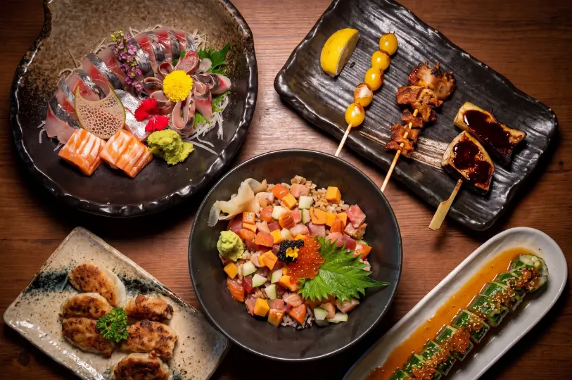 An overhead view of a hearty Japanese feast on a wooden table, including a platter of fresh sashimi, a colorful chirashi rice bowl, pan-fried gyoza, grilled skewers, and a dressed cucumber salad.